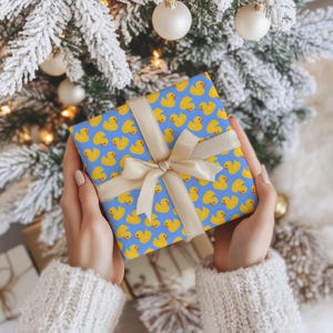 May include: A square gift box wrapped in blue paper featuring a repeating pattern of yellow rubber ducks. The package is tied with a smooth, light beige satin ribbon arranged in a cross pattern and finished with a centred bow on the front.