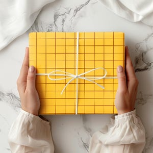 May include: A yellow gift box with a grid pattern and a white ribbon tied in a bow. The box is held by two hands, and the background is a marble surface with a white cloth.