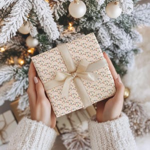 Könnte beinhalten: Ein verpacktes Geschenk mit cremefarbenem Hintergrund und einem Muster aus bunten Lichterketten. Das Geschenk ist mit einem cremefarbenen Band und einer Schleife gebunden. Der Hintergrund umfasst einen Weihnachtsbaum mit weißen Kugeln.