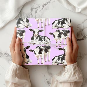 May include: A wrapped gift featuring a repeating pattern of black and white cows on a light purple background. The gift is tied with white string. The overall theme is farm animals.