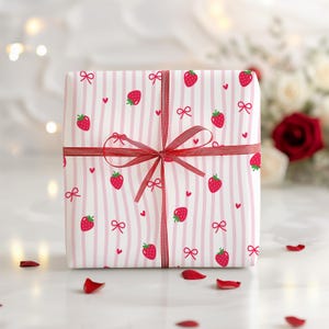 May include: A square gift wrapped in white paper with pink vertical stripes, red strawberries, bows, and hearts. The gift is tied with a red ribbon and bow. Red rose petals are scattered around the gift.
