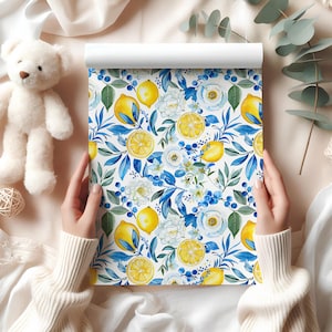 May include: A roll of wallpaper with a watercolour design of lemons, white flowers, and blue leaves on a white backdrop. The lemons are yellow, and the leaves are green and blue. The wallpaper is held by a person wearing a white sweater.