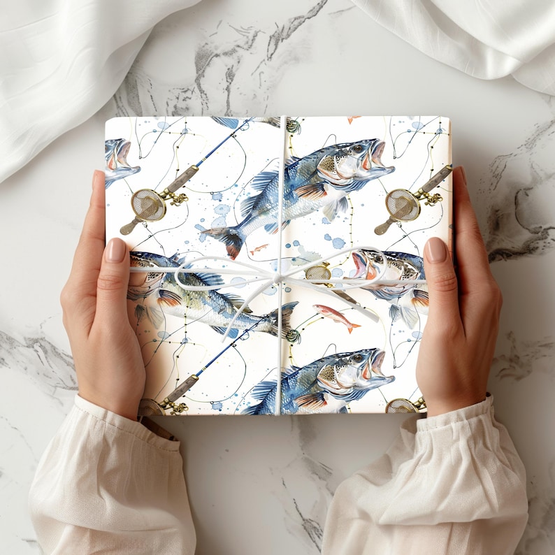 Puede incluir: Una caja de regalo rectangular envuelta en papel blanco con un patr&oacute;n de acuarela repetitivo de peces azules y plateados, ca&ntilde;as de pescar y redes peque&ntilde;as, atada con un cordel blanco fino en un patr&oacute;n de cruz simple.