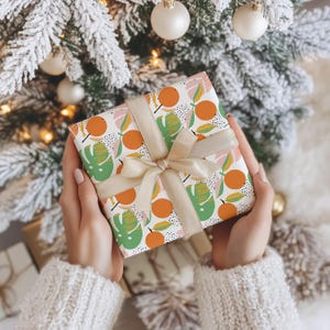 Puede incluir: Una caja de regalo cuadrada envuelta en papel blanco con un patr&oacute;n repetitivo de c&iacute;rculos naranjas y hojas tropicales verdes con detalles de puntos negros. La caja est&aacute; atada con una cinta de sat&eacute;n de color beige claro en un lazo.