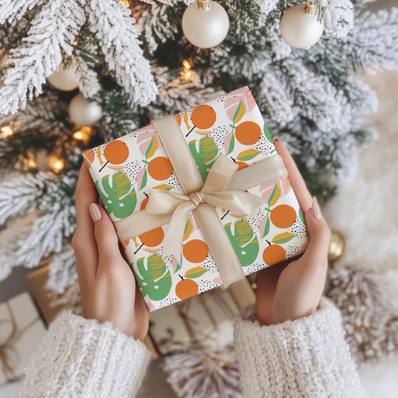 Puede incluir: Una caja de regalo cuadrada envuelta en papel blanco con un patr&oacute;n repetitivo de c&iacute;rculos naranjas y hojas tropicales verdes con detalles de puntos negros. La caja est&aacute; atada con una cinta de sat&eacute;n de color beige claro en un lazo.