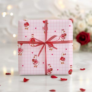May include: A wrapped gift with pink wrapping paper featuring a grid pattern and cartoon geese holding hearts and flowers. The gift is tied with a red ribbon. Red rose petals are scattered around the gift.