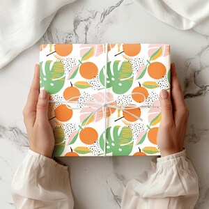 Puede incluir: Una caja de regalo cuadrada envuelta en papel blanco con un patr&oacute;n repetitivo de c&iacute;tricos de color naranja brillante, hojas de monstera verdes y peque&ntilde;os puntos negros, atada con un cordel blanco fino dispuesto en forma de cruz sobre el centro.