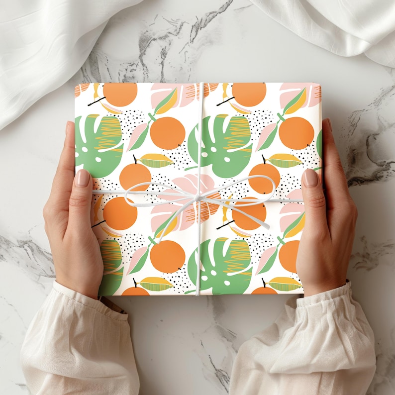 Puede incluir: Una caja de regalo cuadrada envuelta en papel blanco con un patr&oacute;n repetitivo de c&iacute;tricos de color naranja brillante, hojas de monstera verdes y peque&ntilde;os puntos negros, atada con un cordel blanco fino dispuesto en forma de cruz sobre el centro.