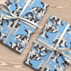 May include: Two gift boxes wrapped in light blue paper featuring a pattern of black and white cows. Each box is tied with a white ribbon. The boxes are on a light brown wooden surface.