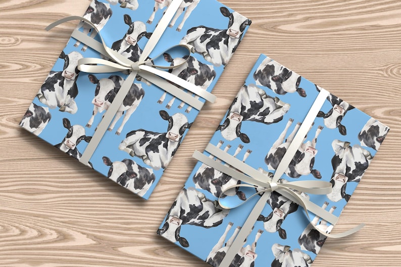 May include: Two gift boxes wrapped in light blue paper featuring a pattern of black and white cows. Each box is tied with a white ribbon. The boxes are on a light brown wooden surface.