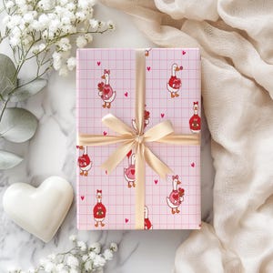 May include: A wrapped gift with pink wrapping paper featuring a pattern of cartoon geese holding flowers and hearts. The gift is tied with a gold ribbon and bow. White flowers and a white heart are in the background.