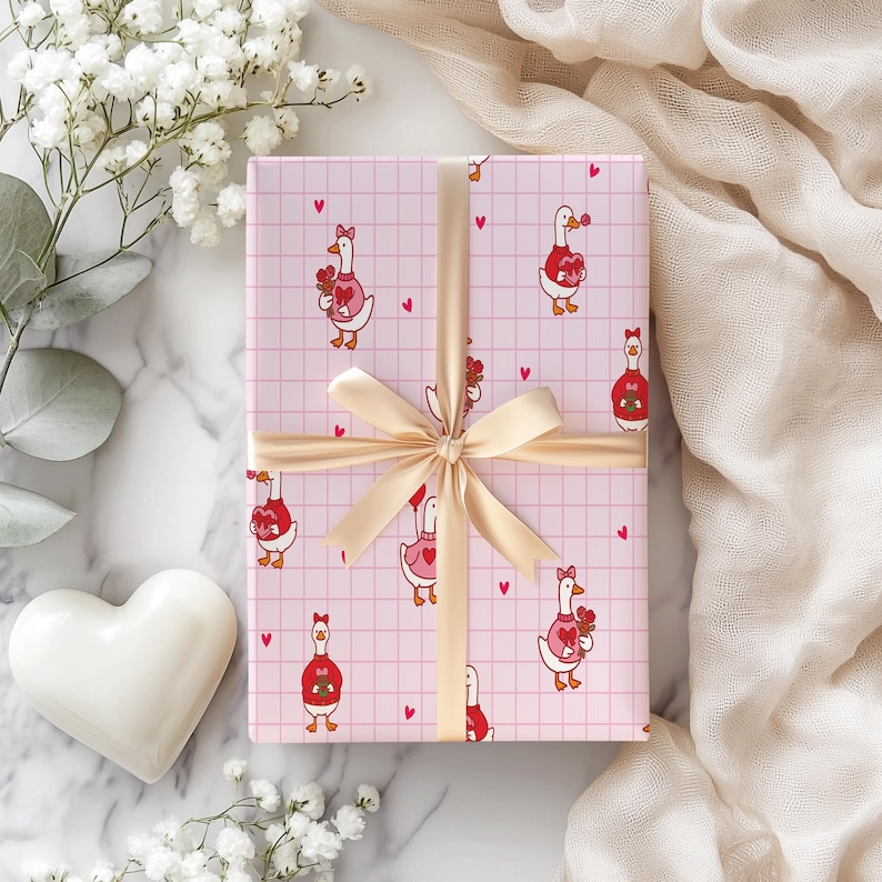 May include: A wrapped gift with pink wrapping paper featuring a pattern of cartoon geese holding flowers and hearts. The gift is tied with a gold ribbon and bow. White flowers and a white heart are in the background.