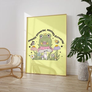 May include: A yellow poster with a green frog sitting on a pink and white mushroom. The frog is wearing a crown and holding a cup. The text on the poster reads "Naturally Introverted, Selectively Extroverted".