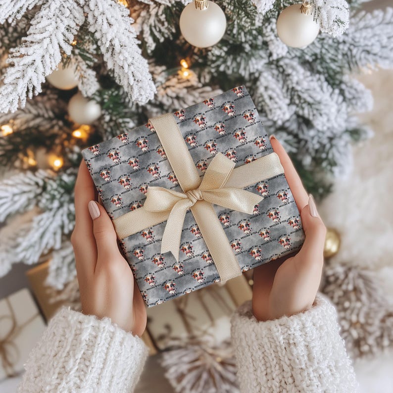 May include: A square gift box wrapped in gray paper featuring a repeating pattern of cow faces and the text "MOO-DY B*TCH" in black lettering. The box is tied with a smooth, cream-colored satin ribbon arranged in a centered bow across the front.