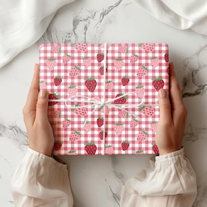 May include: A rectangular gift box wrapped in paper featuring a red and white gingham check pattern decorated with a repeating print of small red and pink strawberries. The package is tied with a thin white string tied in a simple bow.
