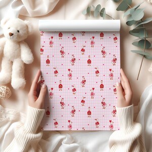 May include: A roll of pink patterned paper featuring cartoon geese holding hearts, set against a grid background. The geese are in red and pink, with red hearts scattered across the surface. A white teddy bear and a ball of twine are in the background.