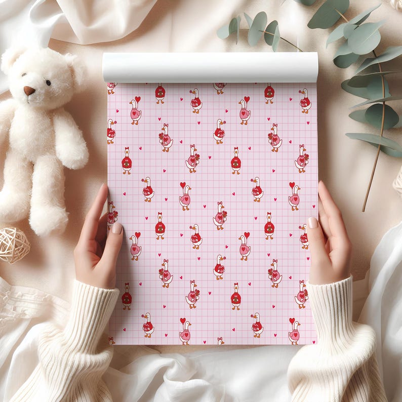 May include: A roll of pink patterned paper featuring cartoon geese holding hearts, set against a grid background. The geese are in red and pink, with red hearts scattered across the surface. A white teddy bear and a ball of twine are in the background.