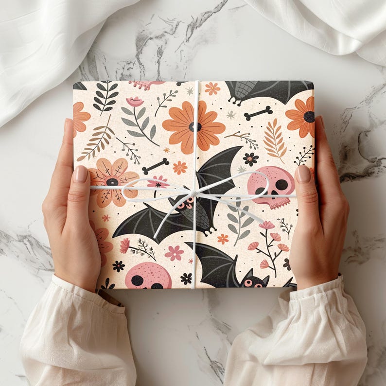 May include: A wrapped gift box featuring a Halloween-themed pattern. The design includes black bats, pink skulls, orange flowers, and bone accents on a cream background. The box is tied with white twine.