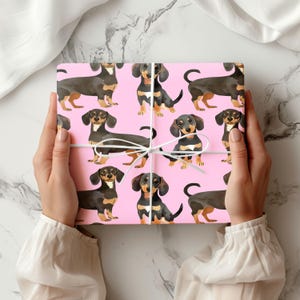 Könnte beinhalten: Rosa Geschenkpapier mit einem sich wiederholenden Muster aus schwarzen und braunen Dackeln. Die Hunde sind in verschiedenen Posen, darunter sitzend, stehend und laufend.