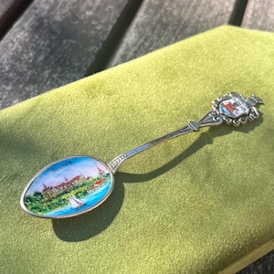 May include: A vintage silver spoon with a colorful enamel scene of a building and lake, labeled "Schloß". The spoon has a decorative handle with a crest. The spoon is resting on a green velvet surface.