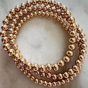 Pode incluir: Três pulseiras douradas com contas. As pulseiras são feitas de pequenas contas redondas e brilhantes de ouro. As contas são enfiadas para formar uma forma circular. As pulseiras são exibidas em uma superfície branca.