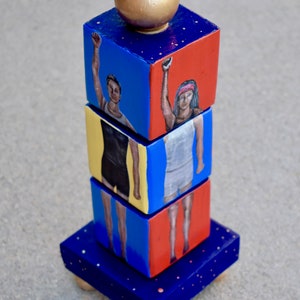 Puede incluir: Una escultura de madera apilada con una esfera dorada en la parte superior. Cada bloque presenta una imagen pintada de una persona con los brazos en alto. Los bloques están pintados en tonos de azul, rojo y amarillo.