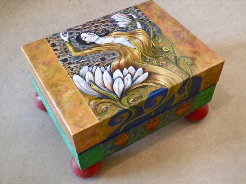 May include: A wooden box with a painted design of a woman in a golden dress standing in a white flower. The box has a blue, green, and orange base with red wooden legs.