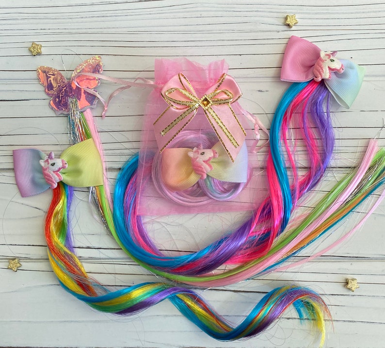 May include: Three colorful hair clips with unicorn charms and rainbow hair extensions. The clips are pink, blue, and green with a white bow and a unicorn charm on each. The hair extensions are a mix of pink, blue, green, and yellow.
