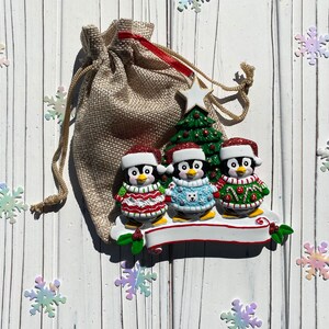May include: A Christmas ornament featuring three penguin figurines wearing Santa hats and sweaters. The penguins stand on a white banner with a green Christmas tree and a burlap gift bag in the background. Iridescent snowflake decorations are scattered around.