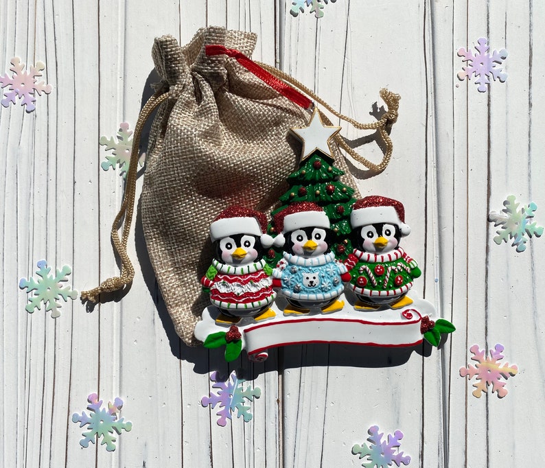 May include: A Christmas ornament featuring three penguin figurines wearing Santa hats and sweaters. The penguins stand on a white banner with a green Christmas tree and a burlap gift bag in the background. Iridescent snowflake decorations are scattered around.