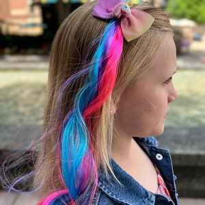 May include: A young person with blonde hair wearing a denim jacket and a colorful hair extension with a unicorn bow clip.