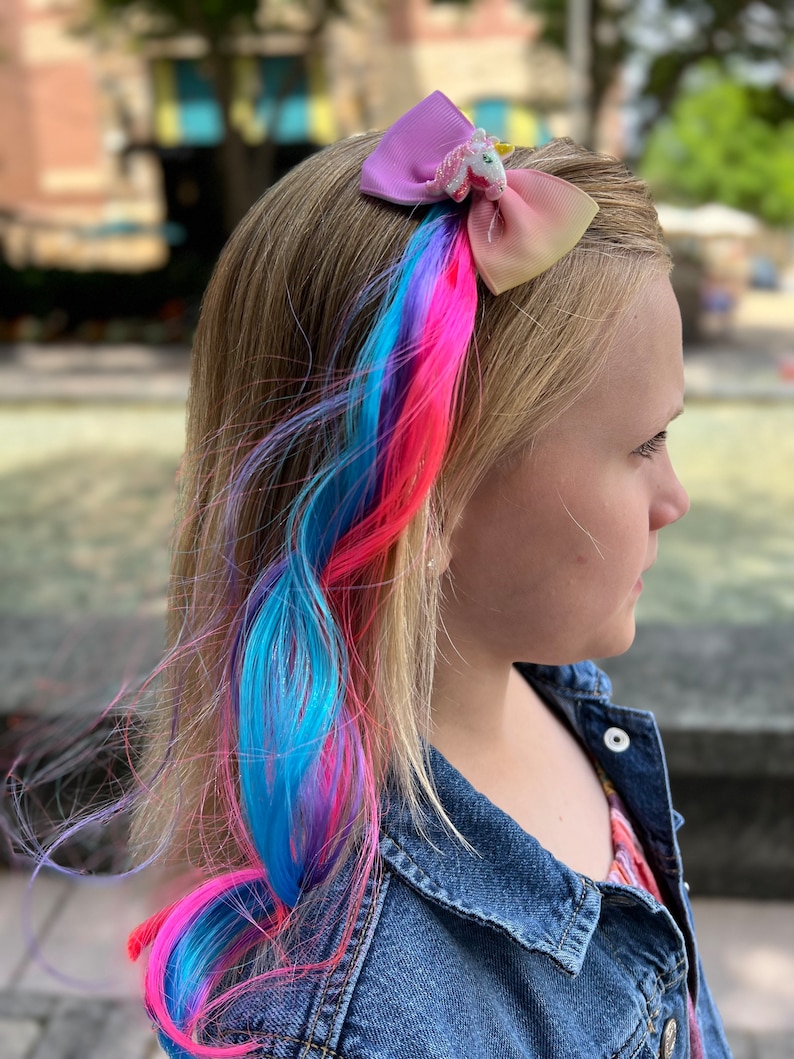 May include: A young person with blonde hair wearing a denim jacket and a colorful hair extension with a unicorn bow clip.