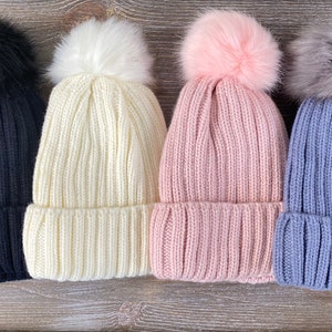 May include: Four knitted winter hats with faux fur pom poms in black, white, pink, and blue.