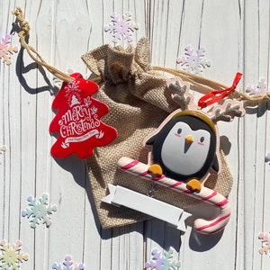 May include: A Christmas-themed arrangement featuring a penguin ornament with a candy cane, a red tree-shaped tag with "Merry Christmas" text, and a burlap bag. The penguin has a black and white body with a yellow beak and antlers. Decorative snowflakes surround the items.