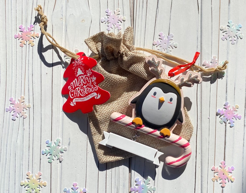 May include: A Christmas-themed arrangement featuring a penguin ornament with a candy cane, a red tree-shaped tag with "Merry Christmas" text, and a burlap bag. The penguin has a black and white body with a yellow beak and antlers. Decorative snowflakes surround the items.