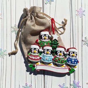 May include: A Christmas ornament featuring a family of penguins wearing Santa hats and sweaters. The penguins are standing on a white banner with space for personalization. A burlap bag and snowflake decorations are in the background.