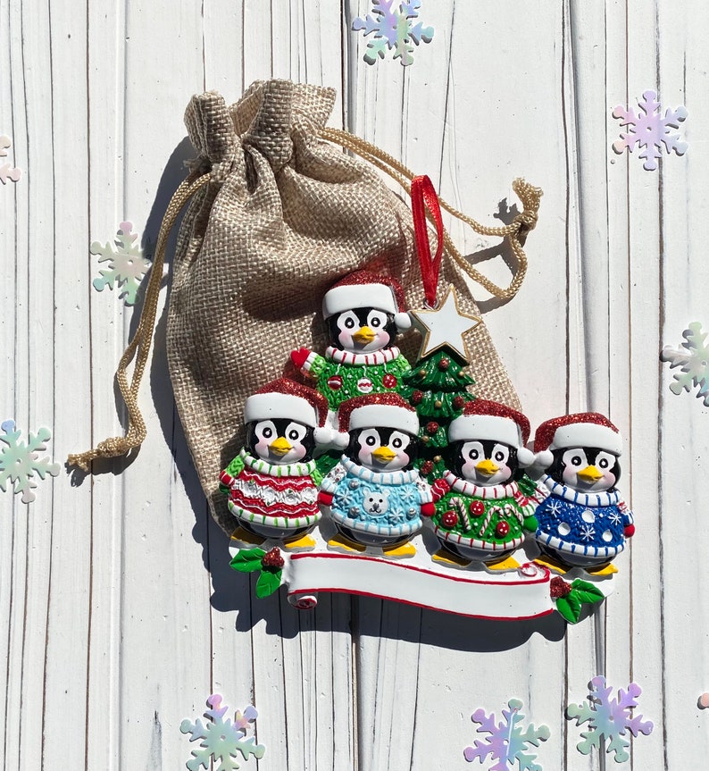 May include: A Christmas ornament featuring a family of penguins wearing Santa hats and sweaters. The penguins are standing on a white banner with space for personalization. A burlap bag and snowflake decorations are in the background.
