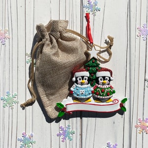 May include: A Christmas ornament featuring two penguins wearing Santa hats and sweaters, standing in front of a Christmas tree. The ornament has a blank banner and is next to a burlap gift bag. Decorative snowflakes are scattered around.