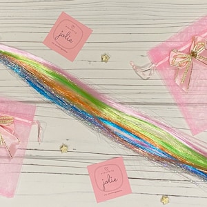 May include: A set of rainbow colored hair extensions with glitter, packaged in two pink organza bags with gold ribbon bows. The bags have a small white square tag with the text "everything jolie" on it.