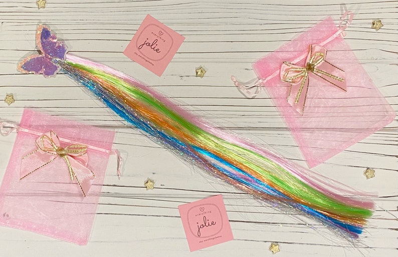 May include: A set of rainbow colored hair extensions with glitter, packaged in two pink organza bags with gold ribbon bows. The bags have a small white square tag with the text "everything jolie" on it.