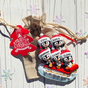 May include: A Christmas ornament featuring a family of penguins on a sled. Each penguin wears a Santa hat and has a name on it. A red tree-shaped tag with "Merry Christmas and Happy New Year" text is attached to a burlap bag.