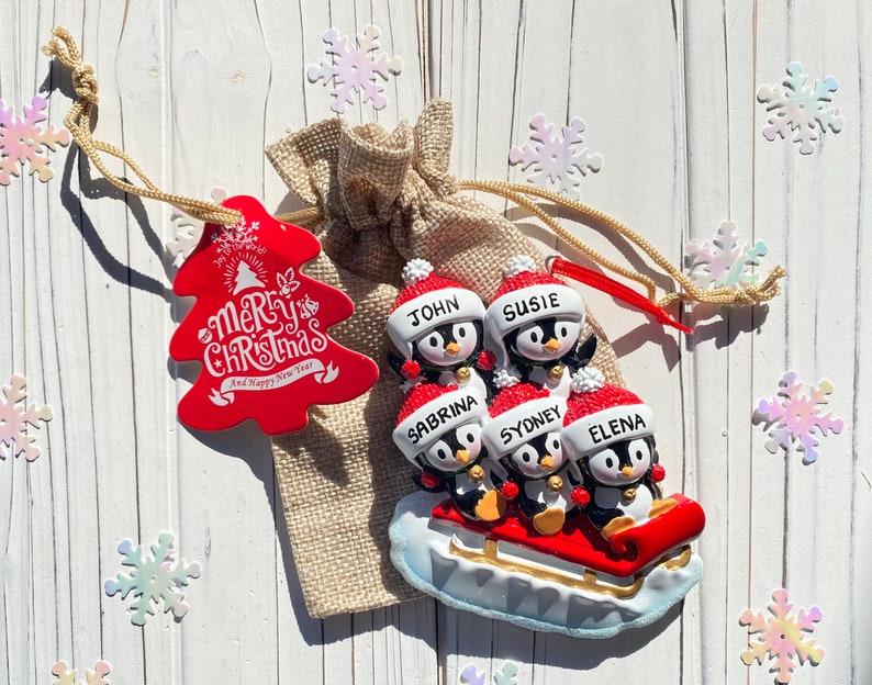 May include: A Christmas ornament featuring a family of penguins on a sled. Each penguin wears a Santa hat and has a name on it. A red tree-shaped tag with "Merry Christmas and Happy New Year" text is attached to a burlap bag.