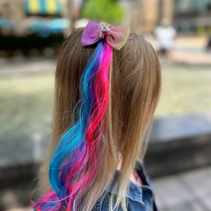 May include: A girl with long blonde hair has a colorful hair extension in a ponytail. The hair extension is blue, pink, and purple. A unicorn-shaped hair bow is holding the ponytail in place.