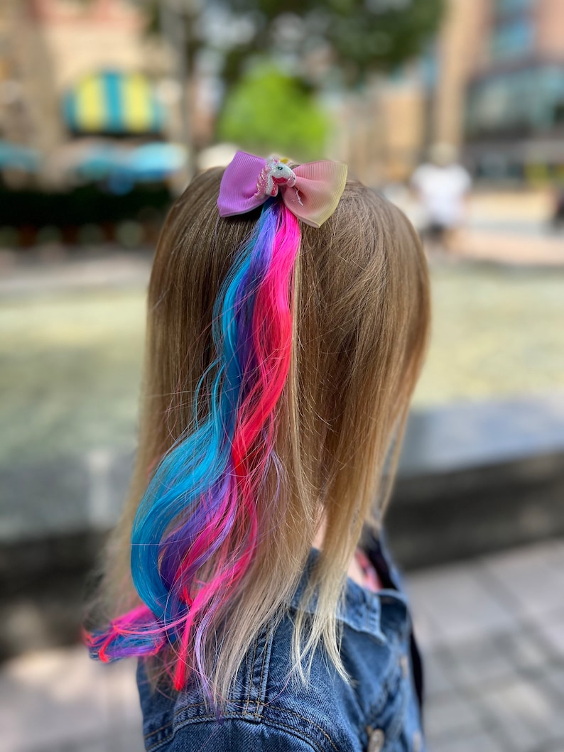 May include: A girl with long blonde hair has a colorful hair extension in a ponytail. The hair extension is blue, pink, and purple. A unicorn-shaped hair bow is holding the ponytail in place.