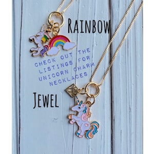 May include: Two gold necklaces with charms. The top necklace has a rainbow and unicorn charm. The bottom necklace has a diamond and unicorn charm. The text "RAINBOW JEWEL" is written on the wooden background. The text "CHECK OUT THE LISTINGS FOR UNICORN CHARM NECKLACES" is also written on the background.