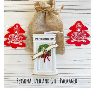 May include: A white door-shaped Christmas ornament with a green wreath and red bow, the address "140 Yosemite Way" and the year "2021". Two red tree-shaped ornaments with "Merry Christmas" text are on either side. A burlap gift bag is behind the ornament.