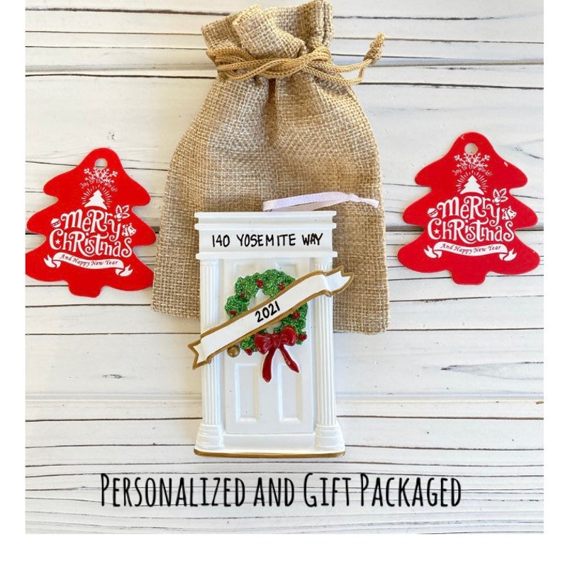 May include: A white door-shaped Christmas ornament with a green wreath and red bow, the address "140 Yosemite Way" and the year "2021". Two red tree-shaped ornaments with "Merry Christmas" text are on either side. A burlap gift bag is behind the ornament.