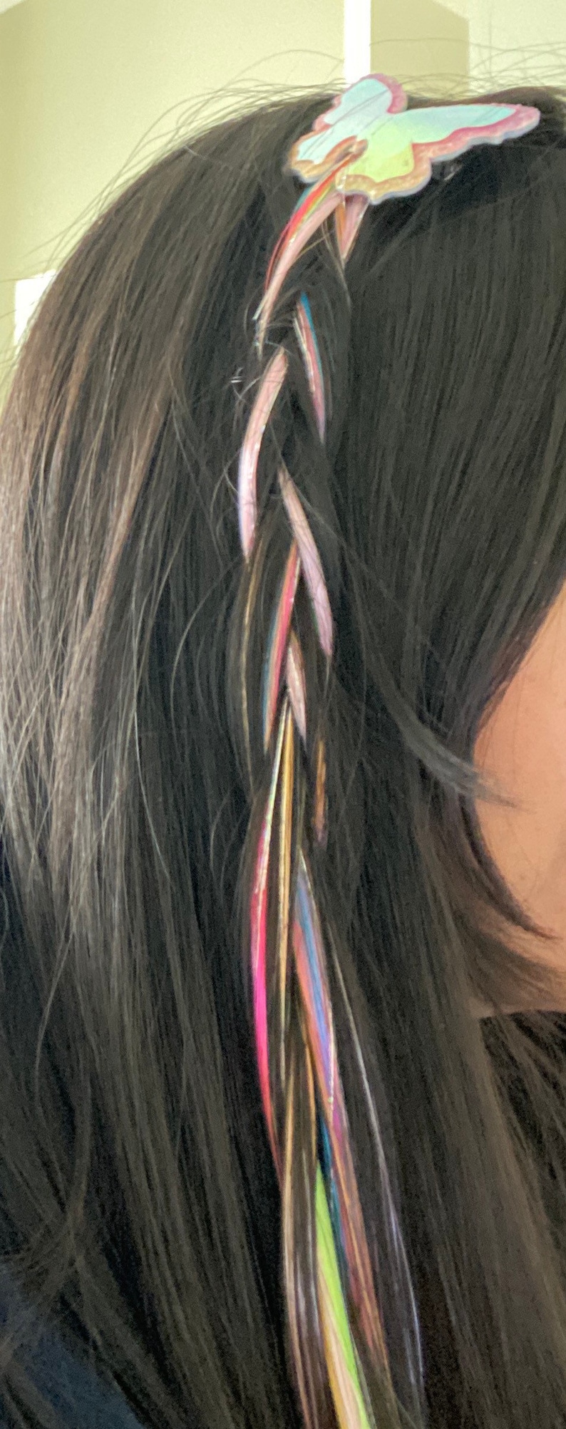 May include: A braided hairstyle with colorful hair extensions and a butterfly-shaped hair clip. The hair extensions are pink, blue, green, yellow, and gold.