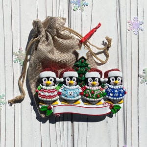 May include: A Christmas ornament featuring four penguin figures wearing festive sweaters and Santa hats. A small Christmas tree and a star are behind the penguins. The ornament is attached to a burlap bag with a red ribbon.
