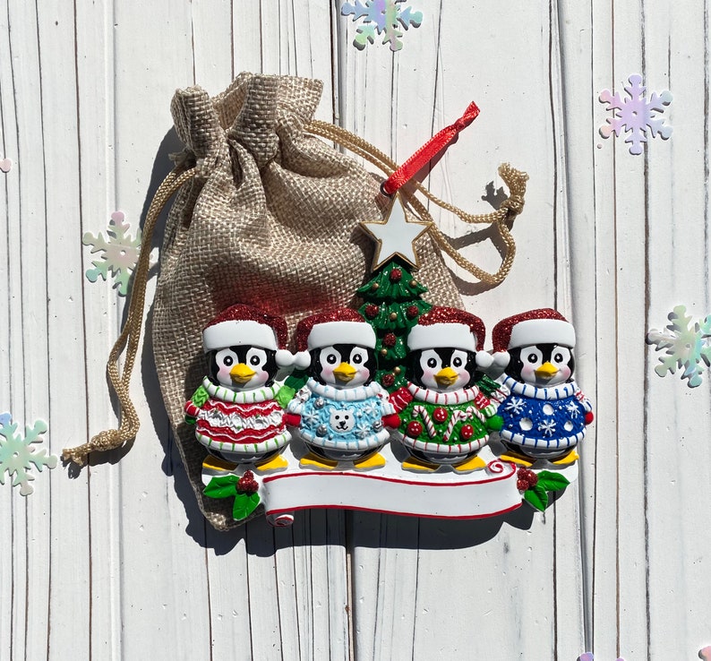 May include: A Christmas ornament featuring four penguin figures wearing festive sweaters and Santa hats. A small Christmas tree and a star are behind the penguins. The ornament is attached to a burlap bag with a red ribbon.